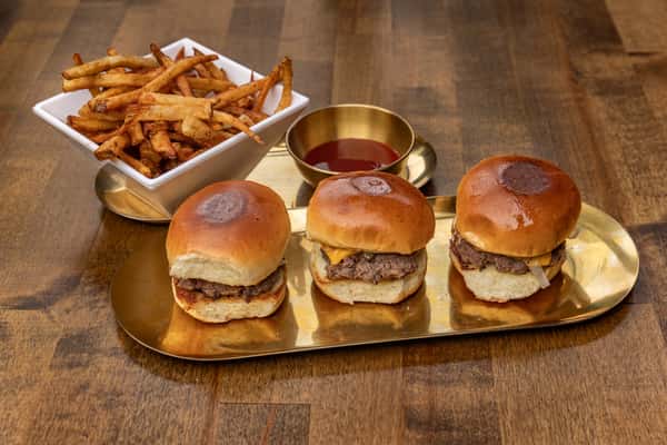 Trio of gourmet beef sliders on brioche buns served on a gold tray, accompanied by seasoned French fries and a side of ketchup in a modern restaurant setting.