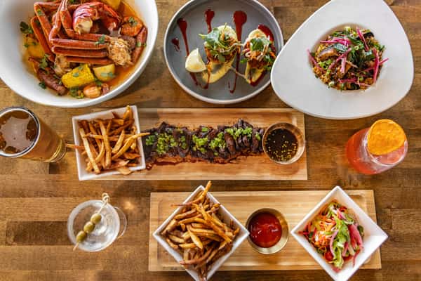 Overhead spread of surf-and-turf dishes including crab legs, tacos, grilled steak with fries, and cocktails in a vibrant modern restaurant setting.