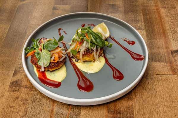 Twin crab cakes on a gray ceramic plate, finished with tangy yellow sauce, pickled slaw, and decorative streaks of house-made red sauce.