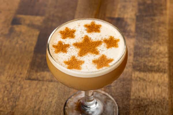 Frothy espresso martini garnished with cinnamon star designs, served in a coupe glass on a wooden table.