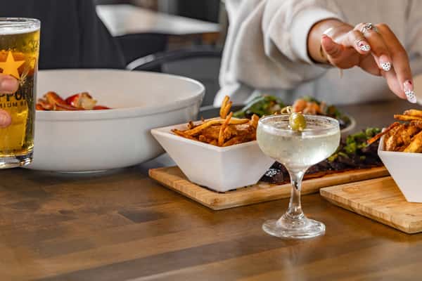 Casual dining setup with seasoned fries, martini garnished with olives, and Sapporo beer, as guests reach for food in a lively restaurant setting.