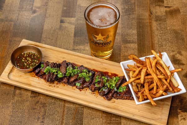 Grilled marinated steak slices topped with chopped scallions, served with golden French fries, spicy dipping sauce, and a pint of Sapporo beer on a wooden platter.