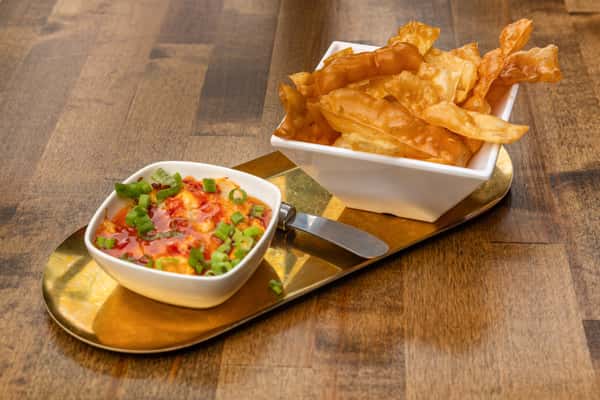 Golden fried wonton chips served with a spicy cheese dip garnished with scallions, presented on a modern gold tray.