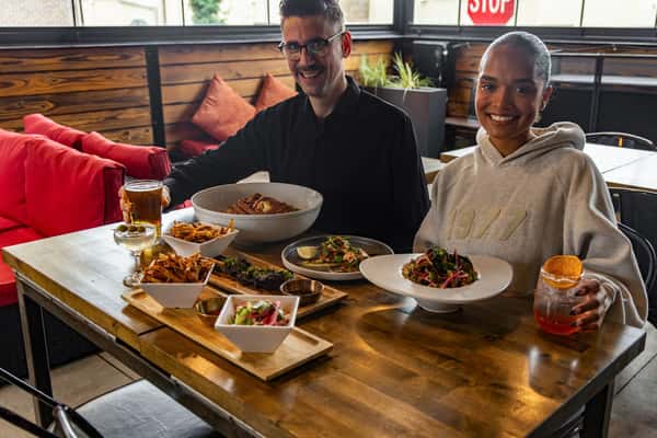 Smiling couple seated at a restaurant table enjoying an Asian fusion spread with crab legs, steak, salad, fries, and craft cocktails.
