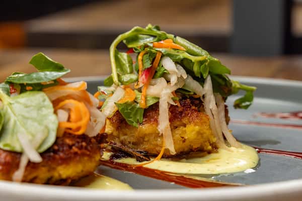Close-up of golden crab cakes topped with pickled vegetables and microgreens, served over creamy aioli and streaks of red sauce on a modern gray plate.