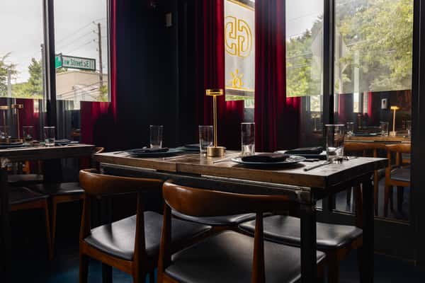 Cozy restaurant setting with wooden tables, leather chairs, and golden table lamps placed on neatly set dining tables beside large windows with deep red curtains.