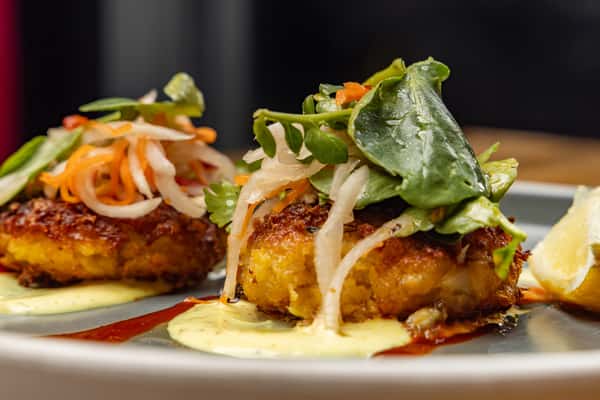 Close-up of crispy crab cakes topped with fresh pickled slaw and greens, plated with lemon and creamy aioli on a slate-gray dish.
