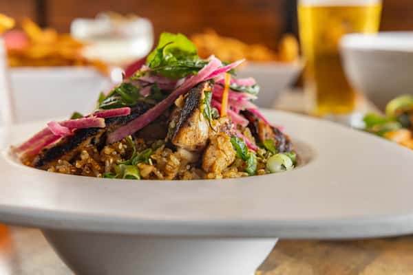 Chargrilled chicken rice bowl topped with pickled onions, fresh herbs, and leafy greens, served in a wide-brimmed white bowl with a craft beer in the background.