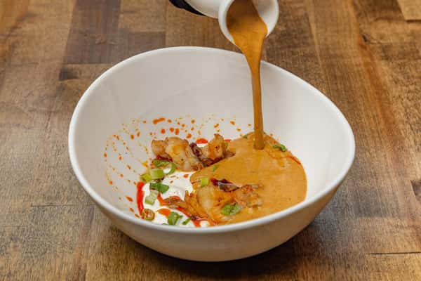 Creamy bisque being poured into a bowl of seared shrimp, scallions, and chili oil, captured mid-action in a white ceramic bowl on a wooden table.