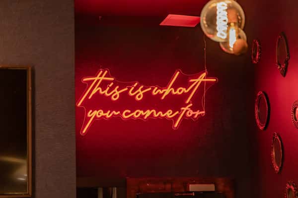 Neon wall sign reading "this is what you come for" in a dimly lit restaurant interior with warm ambiance and vintage mirrors.