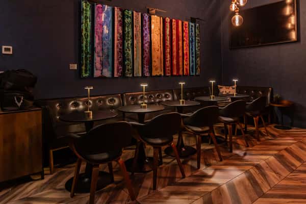Chic restaurant lounge with a long tufted leather banquette, modern black chairs, and individual gold table lamps, accented by a colorful abstract wall installation and warm pendant lighting.