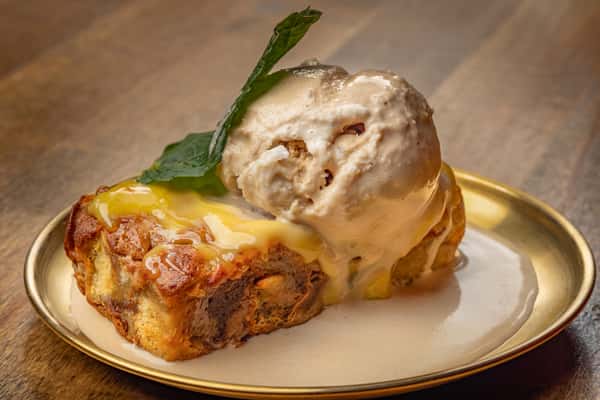 Rich bread pudding topped with melting vanilla ice cream and a drizzle of custard sauce, finished with a mint leaf garnish.