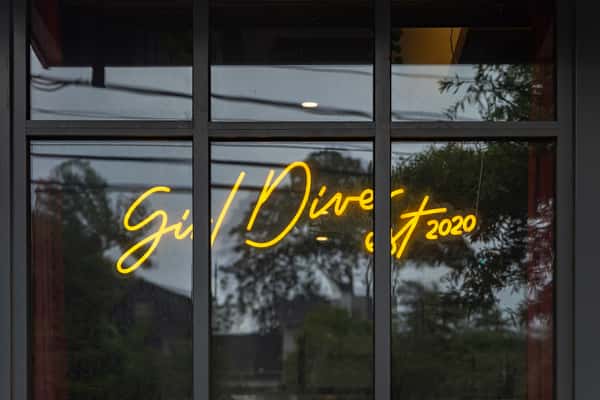 Neon window signage reading "Girl Diver est 2020," glowing against a reflective glass surface with a backdrop of trees and power lines.