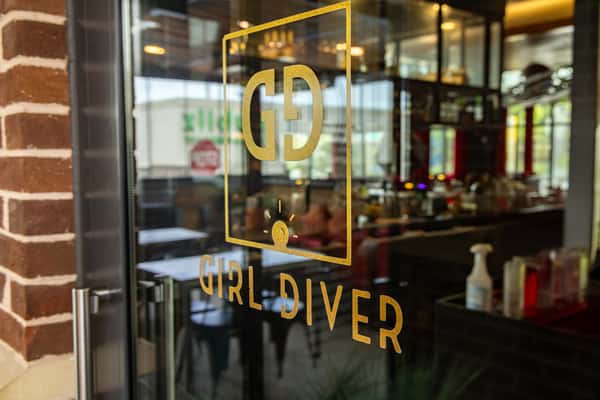 Glass door entrance with golden "Girl Diver" logo decal, offering a glimpse into a modern and stylish restaurant interior.