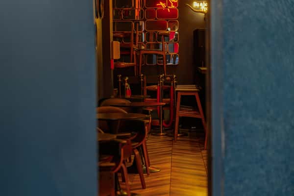 Dimly lit speakeasy-style lounge with black chairs, wooden flooring, gold rope barriers, and red neon signage reflecting in mirrored wall panels.