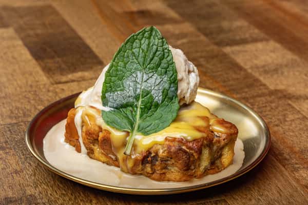Warm bread pudding topped with vanilla ice cream, creamy custard sauce, and a fresh mint leaf, served on a brass-rimmed dessert plate.