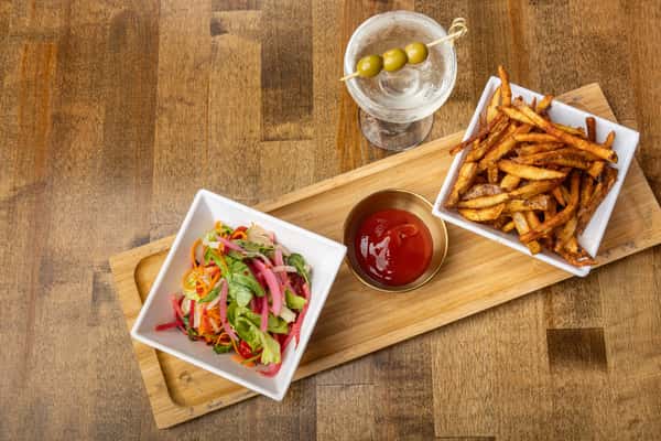 Overhead view of fresh vegetable salad, seasoned French fries, and ketchup, paired with a classic martini with olives on a wooden serving board.