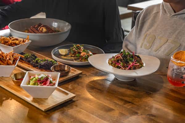 Overhead view of a vibrant table setting with shared dishes including fries, crab cakes, salad, grilled steak, and cocktails in a modern restaurant.