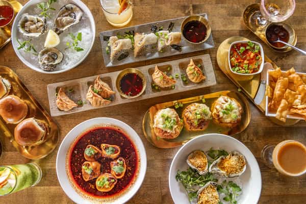 Overhead view of an eclectic mix of Asian fusion dishes including raw oysters, spicy dumplings, crispy spring rolls, sliders, and vibrant cocktails on a rustic wooden table.