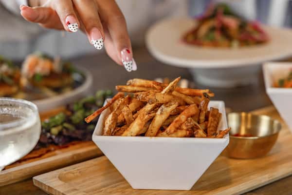 Hand reaching for crispy golden French fries served in a white bowl with a side of ketchup, martini and plated steak in the background.