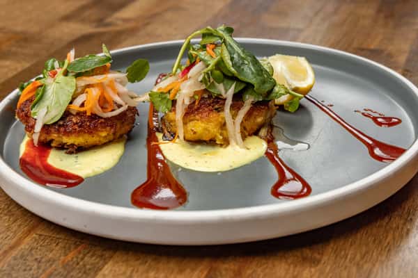 Plated crab cakes topped with pickled vegetables and microgreens, served with tangy aioli, lemon wedge, and artistic streaks of red sauce.