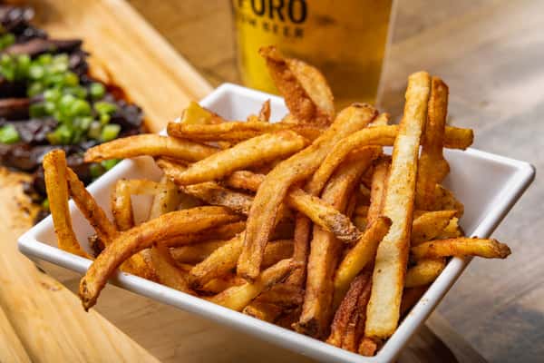 Close-up of crispy hand-cut French fries seasoned to perfection, served in a square white bowl with a pint of beer and sliced steak in the background.