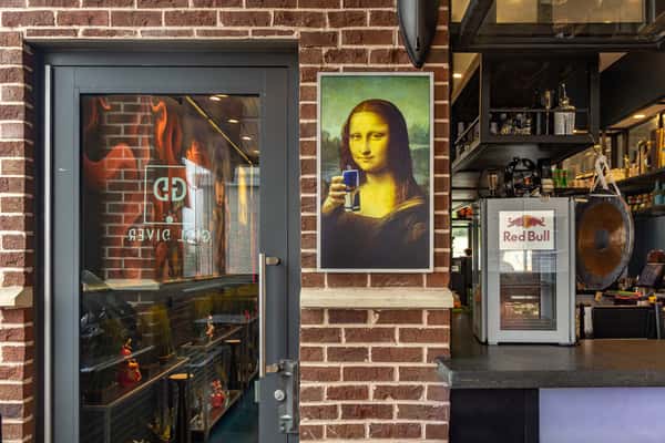 Restaurant interior featuring a playful Mona Lisa artwork holding a Red Bull can, set against an exposed brick wall beside a Red Bull refrigerator and glass entry door.