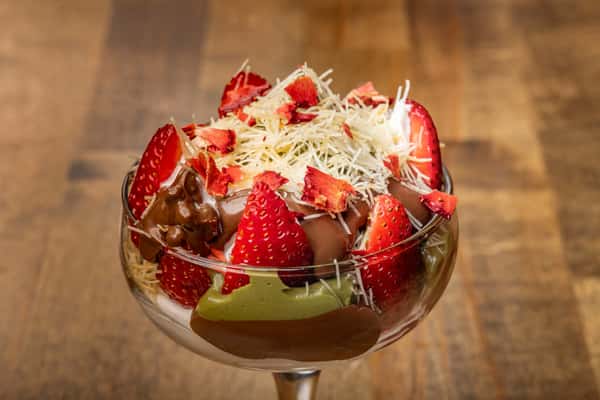 Decadent dessert with layers of green matcha mousse, strawberries, shaved white chocolate, and chocolate drizzle in a clear dessert glass.