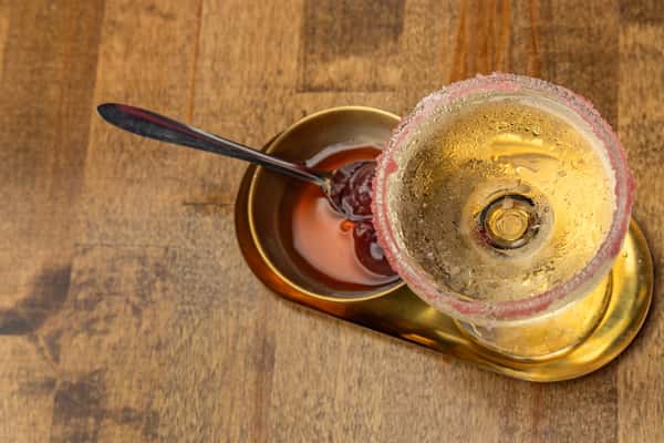 Overhead view of a cocktail with a pink sugar rim beside a brass bowl of cherries in syrup, served on a reflective golden tray.
