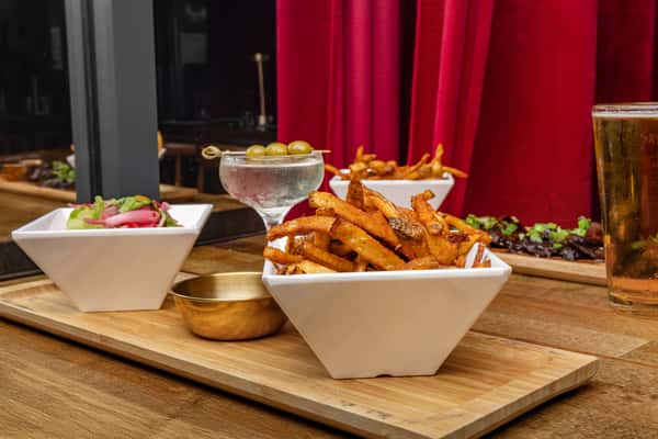 Close-up of crispy golden fries with a side of olives martini, fresh salad, and chargrilled steak bites on a wooden board in a modern dining setting with red curtains.
