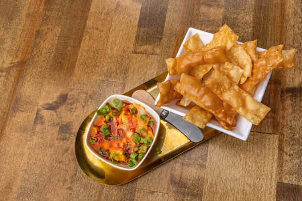Golden fried wonton chips served with spicy melted cheese dip garnished with scallions, plated on a modern gold tray with a cheese knife.