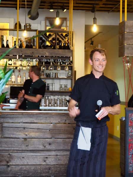 A man in a chef's uniform holding a microphone stands in front of a rustic bar. Another man in a green shirt stands behind the bar.