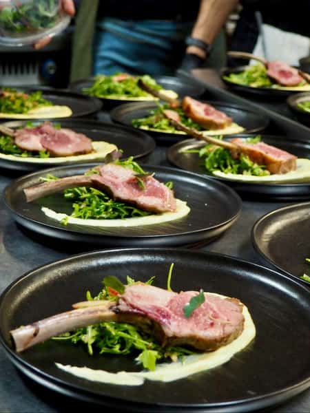 A photo shows lamb chops plated on black dishes in a restaurant. The chops are presented with a small salad and a light sauce.