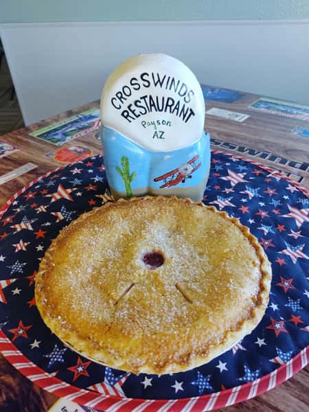 Golden-brown fruit pie with a sugar-crusted top sits on a patriotic-themed placemat at Crosswinds Restaurant in Payson, AZ, with a decorative display in the background.