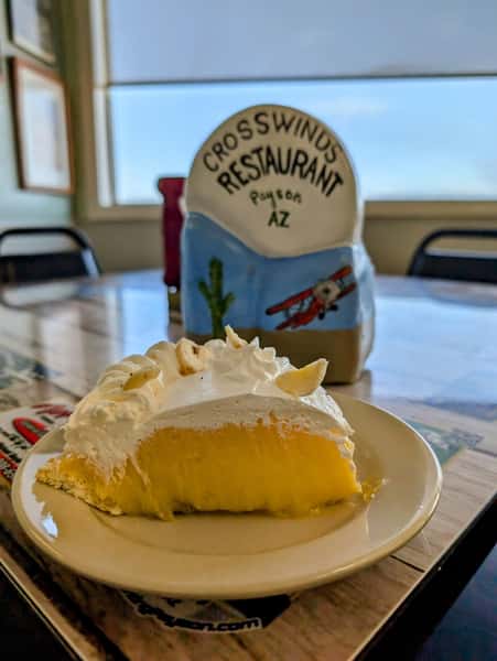 Slice of banana cream pie topped with whipped cream, served at Crosswinds Restaurant in Payson, AZ, with aviation-themed decor in the background.