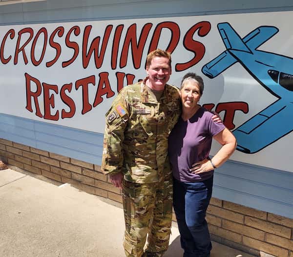 A smiling U.S. Army service member poses with a woman in front of the Crosswinds Restaurant mural in Payson, AZ, featuring bold red text and a blue airplane illustration.