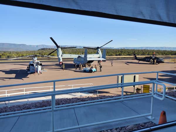 "View of military aircraft, including a V-22 Osprey and helicopters, parked on the Sedona Airport tarmac with personnel nearby, set against a backdrop of red rock mesas and clear blue skies.