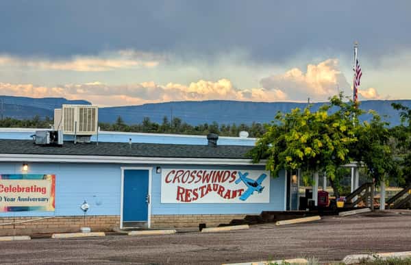 Exterior view of Crosswinds Restaurant in Payson, Arizona, featuring a bright blue building with a vintage airplane mural, set against a dramatic mountain backdrop under cloudy skies.