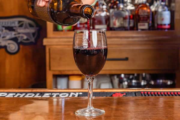 Red wine being poured into a glass at a bar with liquor bottles in the background.
