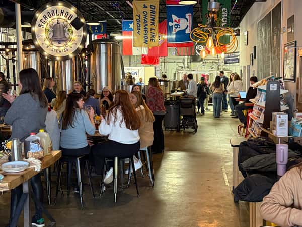 The Schoolhouse Brewing taproom filling up with guests at tables and barstools, with the brewhouse tanks and lit bell sign in the background
