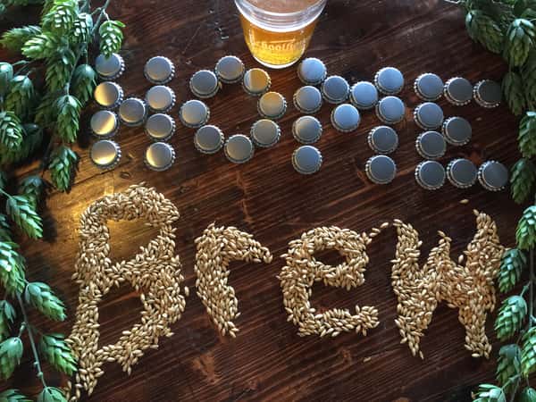 Malted barley spelling the word Brew on a dark wood table, surrounded by hop vines, bottle caps, and a glass of beer