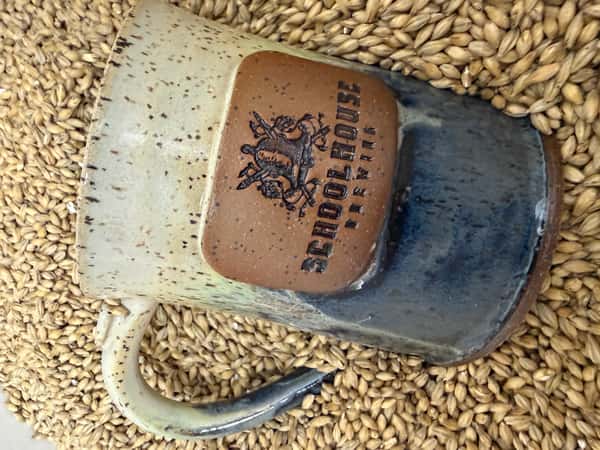 A Schoolhouse Brewing ceramic mug resting in a pile of malted barley from the Schoolhouse homebrew supply shop