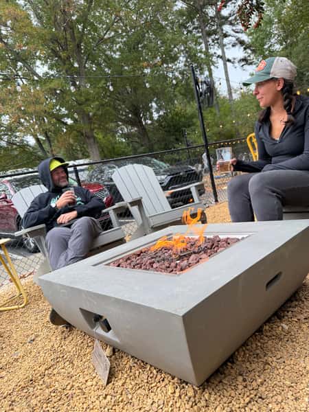 Two guests relaxing in Adirondack chairs with beers around a lit gas fire pit in the Schoolhouse Brewing biergarten