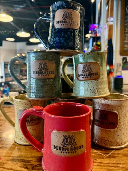 A stack of colorful handmade Schoolhouse Brewing ceramic mugs in blue, green, cream, and red on a wooden bar