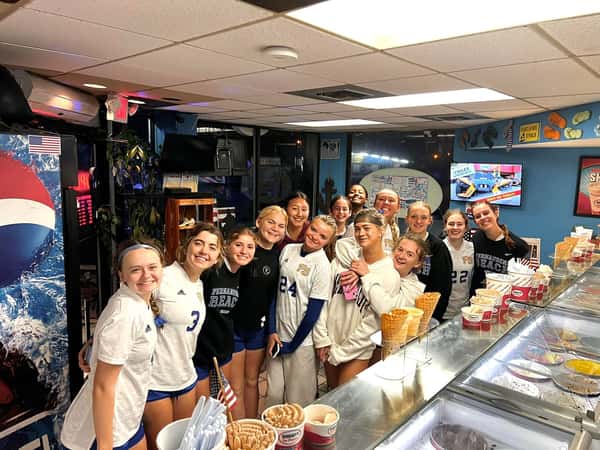 Smiling girls' soccer team gathered inside an ice cream shop, standing by the counter filled with cones and toppings.