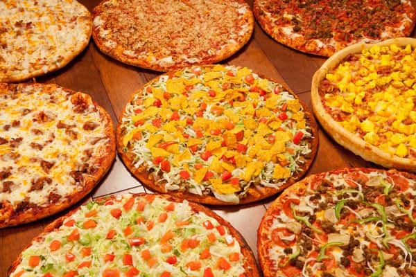 Assorted specialty pizzas on a wooden table, including taco pizza with nacho chips, veggie, meat lovers, and breakfast varieties.