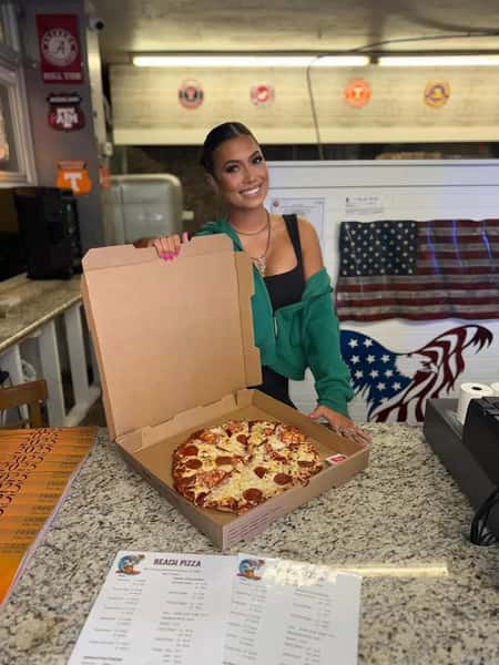Smiling woman behind a counter holds an open pizza box with a fresh pepperoni pizza, standing next to a Beach Pizza menu.