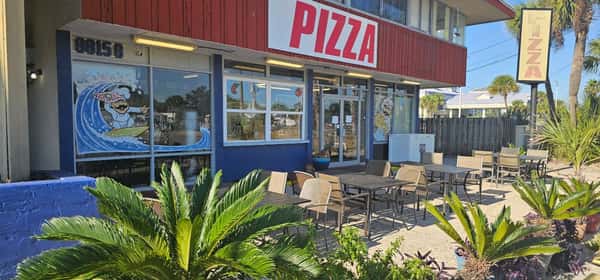 Front patio of Beach Pizza with outdoor seating, palm plants, and large window art featuring a surfing pizza chef.