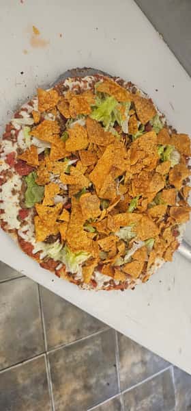 Taco pizza topped with ground beef, lettuce, shredded cheese, and crushed nacho chips on a prep counter.
