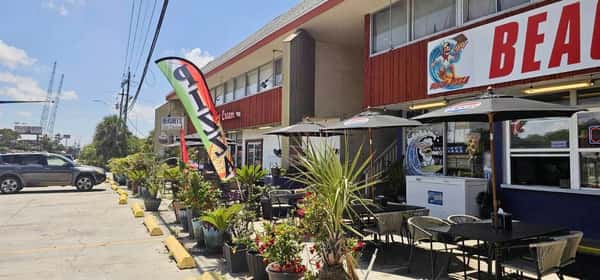 Outdoor view of Beach Pizza restaurant with patio seating, umbrellas, potted plants, and colorful flags along the sidewalk.